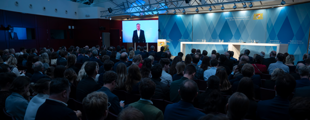 Blick in einen vollbesetzten Saal. Auf einer Leinwand ist Staatsminister Christian Bernreiter zu sehen, der ein Grußwort hält.