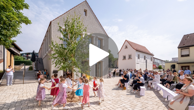 Zur Einweihung der „Neuen Mitte“ in Niederwerrn Ende Juni 2024 waren viele Festgäste gekommen und die Kinder vom Kindergarten tanzten dazu. Im Vordergrund ist eine Schaltfläche „Play“ zu sehen. Wenn man auf das Bild klickt, öffnet sich eine Internetseite von SW-N.TV mit einem Kurzfilm anlässlich der Grundsteinlegung des Projekts in Niederwerrn.