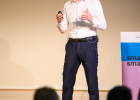Ein Mann steht auf einer Bühne und hält einen Vortrag. Im Hintergrund ist eine Präsentation auf der Leinwand zu sehen. 